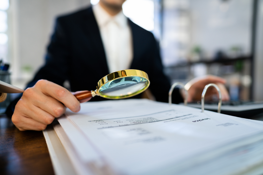 Man sitting at desk has a magnifying glass in his right hand, looking intently at documents. 