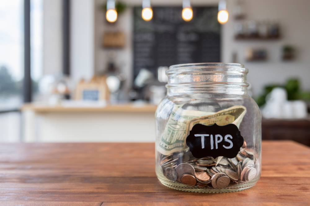 A glass tip jar with American coins and dollar bills sits on a wood countertop in a coffee shop with the background blurred out.